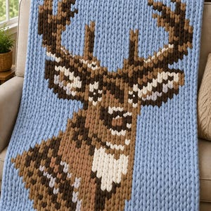 May include: A chunky knit throw blanket featuring a large pixelated deer head design. The blanket is primarily light blue with the deer rendered in shades of brown, tan, and white yarn. The thick, textured stitches create a distinct pattern.