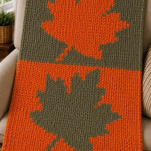 Op de afbeelding: Een grof gebreide plaid met een tweekleurig ontwerp, voorzien van een groot oranje esdoornbladpatroon op een olijfgroene achtergrond in de bovenste helft en een groot olijfgroen esdoornbladpatroon op een oranje achtergrond in de onderste helft.