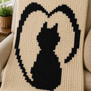 May include: A chunky knit throw blanket in cream yarn featuring a large, centered black silhouette of a sitting cat. The cat is framed by a black heart-shaped outline created with the same thick, textured yarn stitches as the rest of the blanket.