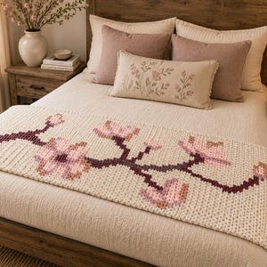 Puede incluir: Un camino de cama de punto color crema con un patrón de ramas horizontales con tallos de color granate oscuro y flores de cerezo de color rosa claro. El camino está colocado a los pies de una cama con ropa de cama crema y cojines decorativos.