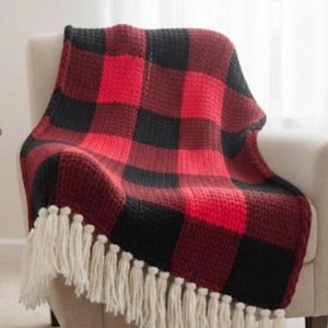 May include: A crocheted throw blanket featuring a red and black buffalo plaid pattern. The blanket is constructed with a textured stitch and is finished with long, cream-colored fringe tassels along the bottom edge of the fabric.