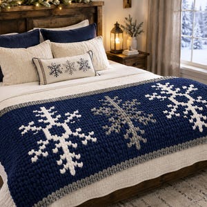 May include: A chunky knit bed runner in deep navy blue featuring three large, textured snowflake motifs in white and light gray. The runner has a thick, braided border in light gray and is draped across a white bedspread with matching pillows.