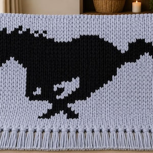 May include: A light blue chunky knit throw blanket featuring a large black horse silhouette in the center. The blanket is constructed with thick yarn stitches and is finished with a row of matching light blue fringe along the bottom edge.
