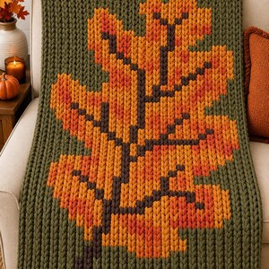 May include: A chunky knit throw blanket featuring a large, central oak leaf design in shades of orange, yellow, and red against a solid olive green background. The blanket is constructed with thick, visible yarn stitches in a vertical ribbed pattern.