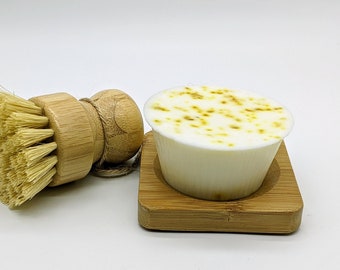 Puede incluir: Una pastilla de jabón blanca con la parte superior moteada de amarillo se encuentra sobre una pequeña bandeja de bambú. También se ve un cepillo de madera con cerdas naturales.