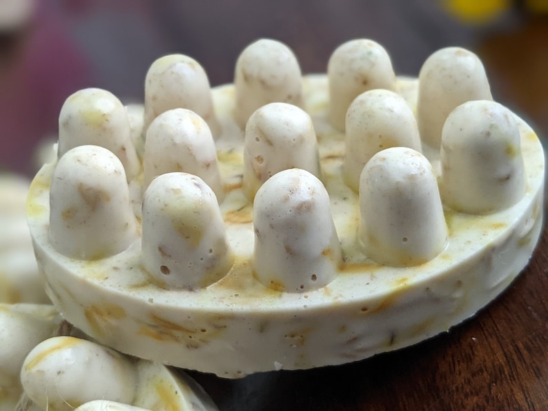 May include: Close-up of a round, off-white soap bar with a textured surface. The soap features numerous rounded, cylindrical protrusions on top. The soap has a speckled appearance with visible flecks of yellow and brown.