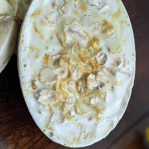 May include: Oval-shaped handmade soap with a creamy white base, speckled with visible oats and other natural ingredients. The soap has a textured surface and a light, natural color palette, suggesting a focus on gentle cleansing and natural skincare.