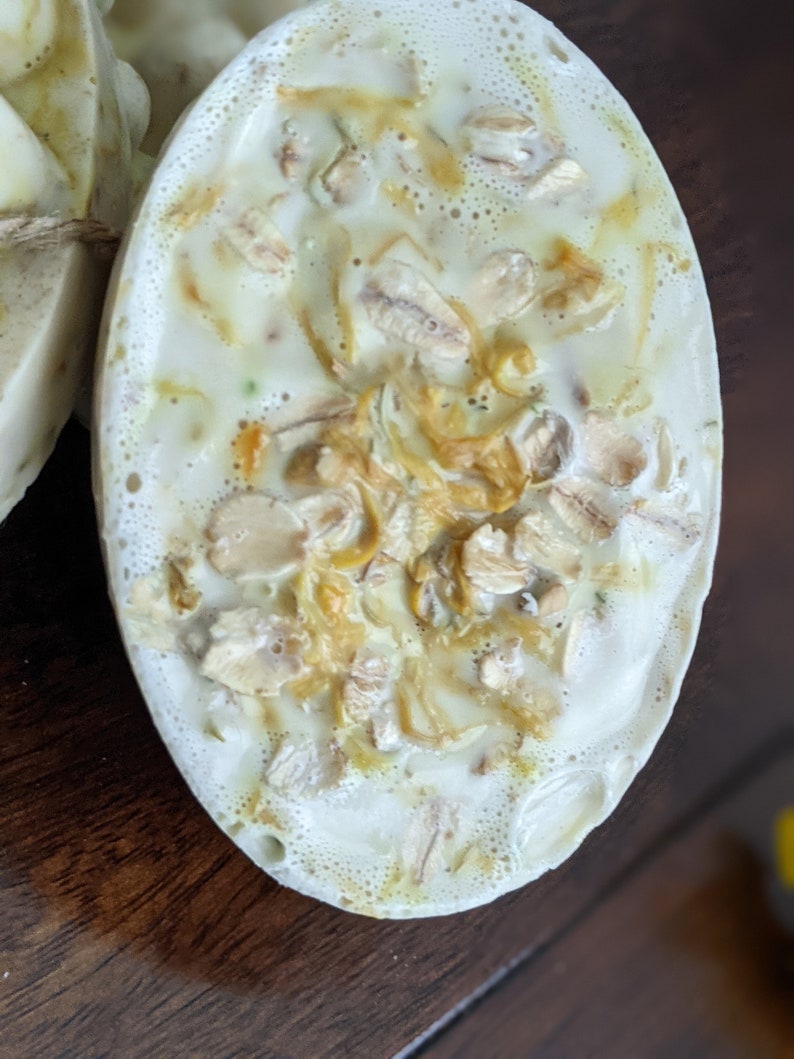 May include: Oval-shaped handmade soap with a creamy white base, speckled with visible oats and other natural ingredients. The soap has a textured surface and a light, natural color palette, suggesting a focus on gentle cleansing and natural skincare.