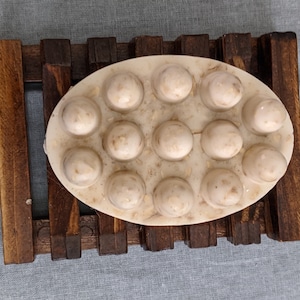 May include: Oval-shaped soap with raised, circular bumps, resting on a wooden soap dish. The soap is off-white with visible flecks, and the dish is made of dark brown wood. The background is a light blue fabric.