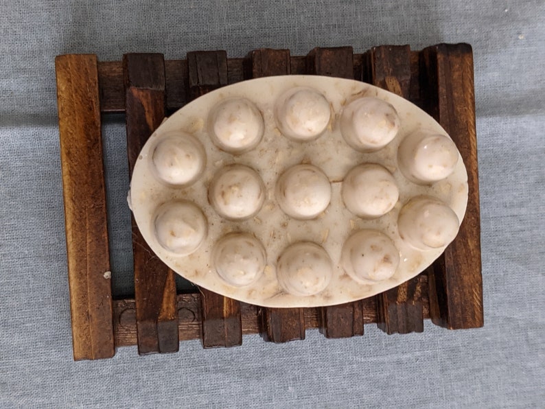May include: Oval-shaped soap with raised, circular bumps, resting on a wooden soap dish. The soap is off-white with visible flecks, and the dish is made of dark brown wood. The background is a light blue fabric.