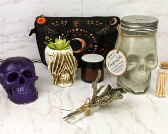 May include: A collection of gothic-themed items, including a purple skull candle, a skull-shaped candle, a black cosmetic bag with celestial designs, a skull-shaped candle, a jar candle, a small jar of matches, and a bundle of herbs.