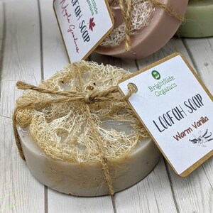 Może przedstawiać: Zestaw trzech mydeł z gąbki lufa, przewiązanych brązowym sznurkiem. Mydła są oznaczone jako "Loofah Soap" i "Warm Vanilla". Mydła leżą na białym drewnianym podłożu.