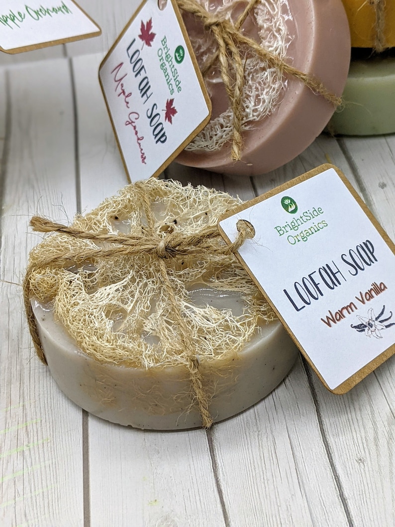Może przedstawiać: Zestaw trzech mydeł z gąbki lufa, przewiązanych brązowym sznurkiem. Mydła są oznaczone jako "Loofah Soap" i "Warm Vanilla". Mydła leżą na białym drewnianym podłożu.