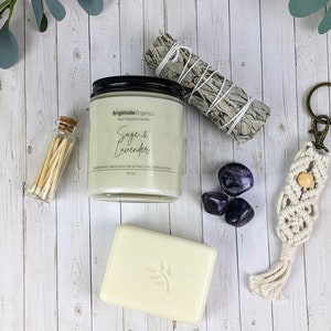 May include: A gift set for relaxation and self-care featuring a soy candle with a sage and lavender scent, a small bundle of white sage, a bar of soap, a small bottle of matches, and three amethyst stones.