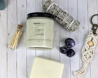 May include: A gift set for relaxation and self-care featuring a soy candle with a sage and lavender scent, a small bundle of white sage, a bar of soap, a small bottle of matches, and three amethyst stones.