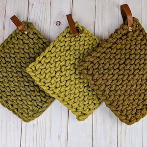 May include: Three green knitted pot holders with brown leather loops. The pot holders are made of thick, textured yarn and have a gradient of green shades.