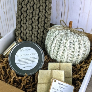 May include: A white gift box filled with brown shredded paper. The box contains a small metal tin candle with the label "Warm Vanilla", two bars of goat milk soap with oatmeal, a knitted white pumpkin, and a brown knitted scarf.