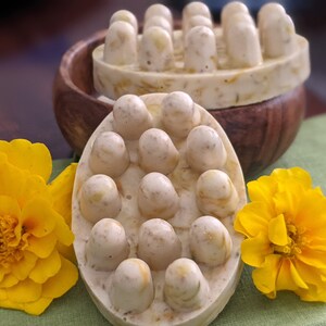 May include: Two handmade massage soaps with rounded knobs for muscle relief. The soaps are light beige with yellow flecks, and are displayed with bright yellow flowers and a wooden bowl.