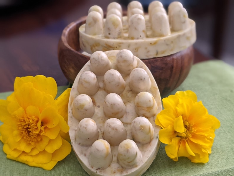 May include: Two handmade massage soaps with rounded knobs for muscle relief. The soaps are light beige with yellow flecks, and are displayed with bright yellow flowers and a wooden bowl.