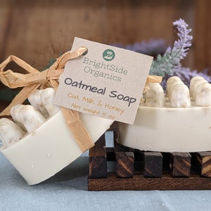 May include: Two bars of oatmeal soap with a tag that reads "BrightSide Organics Oatmeal Soap Oat, Milk & Honey Net weight 5 oz." The soap is light beige with a textured top. One bar is on a wooden soap dish.