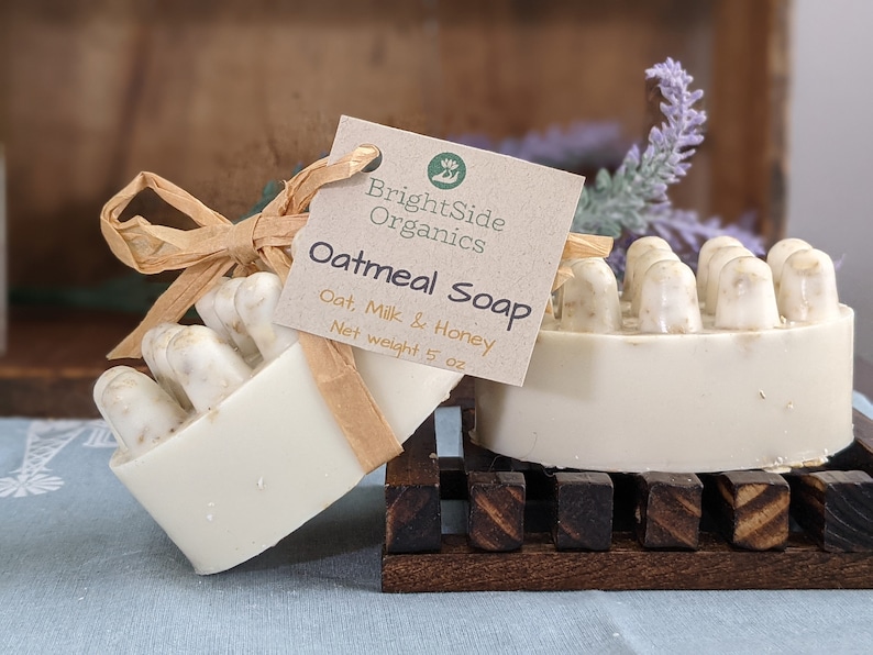 May include: Two bars of oatmeal soap with a tag that reads "BrightSide Organics Oatmeal Soap Oat, Milk & Honey Net weight 5 oz." The soap is light beige with a textured top. One bar is on a wooden soap dish.