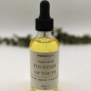 May include: A clear glass bottle with a black dropper top filled with a yellow liquid. The label reads "Brightside Organics Organic Face Oil Fountain of Youth Castor Oil - Jojoba Oil - Frankincense Oil Debary, FL 2 oz."
