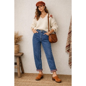 May include: An outfit comprising a cream-coloured V-neck sweater with floral embroidery, blue jeans with plaid cuffs, a brown leather bag, clogs, and a brown cap. The ensemble suggests a casual, autumnal style.