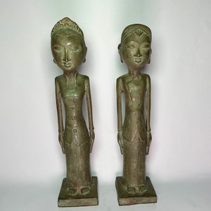 Puede incluir: Dos estatuas de bronce verde de personas de pie con las manos juntas frente a ellas. Las estatuas llevan ropa tradicional y tienen rasgos faciales detallados.