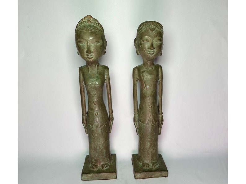 Puede incluir: Dos estatuas de bronce verde de personas de pie con las manos juntas frente a ellas. Las estatuas llevan ropa tradicional y tienen rasgos faciales detallados.