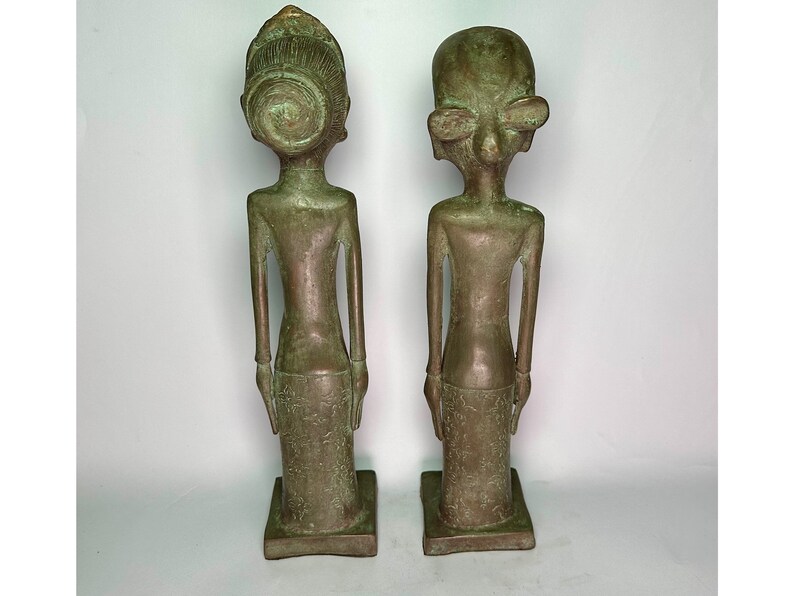 Puede incluir: Dos estatuas de bronce de personas, una con una corona y la otra con ojos grandes. Las estatuas est&aacute;n de pie sobre una superficie blanca.