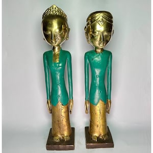 Könnte beinhalten: Zwei goldbemalte Holzstatuen von Menschen in grüner Kleidung. Die Statuen stehen auf einer weißen Oberfläche.