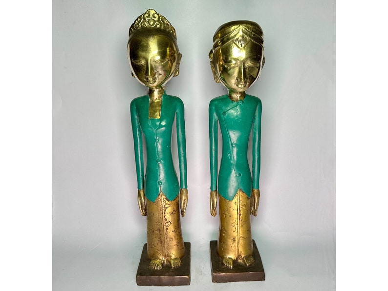 Puede incluir: Dos estatuas de madera pintadas de oro que representan a personas vestidas con ropa verde. Las estatuas est&aacute;n de pie sobre una superficie blanca.