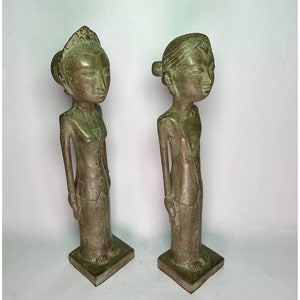 Puede incluir: Dos estatuas de bronce verde de personas de pie una al lado de la otra. Las estatuas son estilizadas y tienen rasgos simples. La estatua de la izquierda lleva una corona y un vestido largo. La estatua de la derecha lleva un tocado sencillo y una prenda larga.