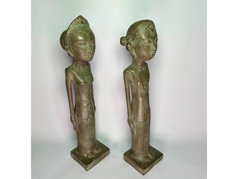 Puede incluir: Dos estatuas de bronce verde de personas de pie una al lado de la otra. Las estatuas son estilizadas y tienen rasgos simples. La estatua de la izquierda lleva una corona y un vestido largo. La estatua de la derecha lleva un tocado sencillo y una prenda larga.