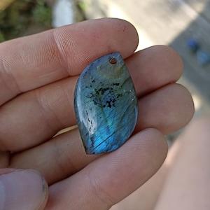 May include: A teardrop-shaped pendant made of iridescent labradorite gemstone with a hole at the top for a chain or cord. The stone has a blue, green, and black color with a shimmery effect.