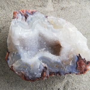 May include: A natural geode stone with a cavity. The stone features layers of white, gray, and blue hues, with reddish-brown edges. The geode is set against a sandy background, showcasing its natural beauty.