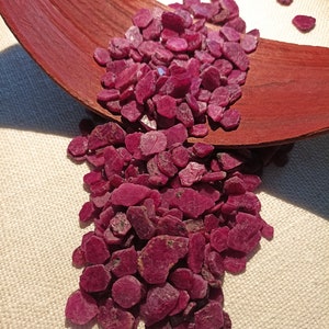 May include: A collection of raw, deep red ruby gemstones. The gemstones are arranged on a dark brown, curved wooden surface, with some spilling over. The background is a light beige fabric.