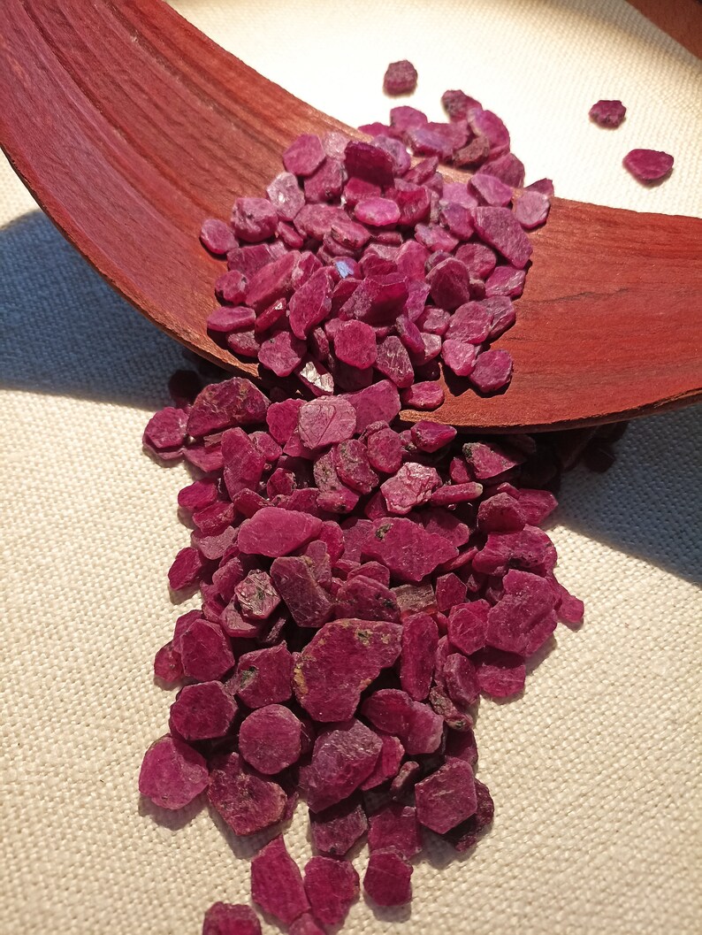 May include: A collection of raw, deep red ruby gemstones. The gemstones are arranged on a dark brown, curved wooden surface, with some spilling over. The background is a light beige fabric.