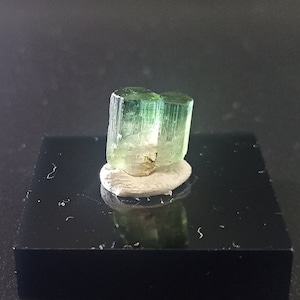 May include: A light green tourmaline crystal specimen. The crystal is a translucent, elongated shape with a vertical orientation. It is mounted on a small, off-white base, which is set on a black, glossy display block. The crystal has a natural, unpolished appearance.