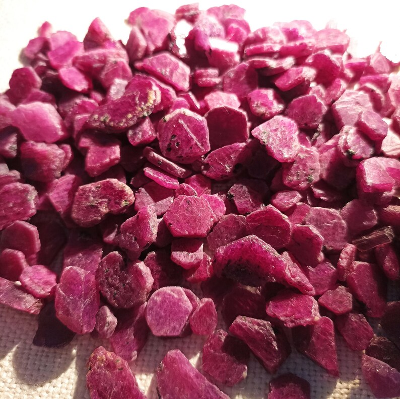 May include: A collection of rough ruby gemstones. The stones are a deep red hue, with a slightly textured surface. The gemstones vary in size and shape, with some appearing more angular and others more rounded. The overall effect is a collection of natural, unpolished rubies.