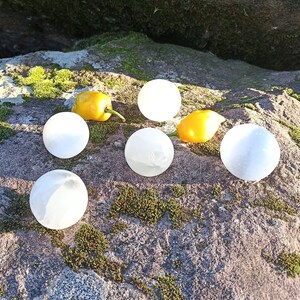 May include: Seven white selenite spheres on a mossy rock surface. The spheres are smooth and polished, with a translucent white color.