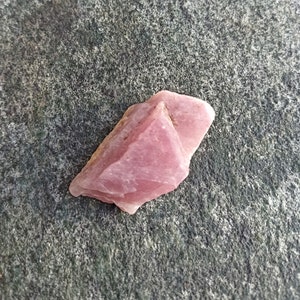 May include: A pink, rough-cut gemstone with a pointed tip, sitting on a gray, textured surface.