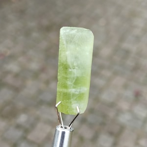 May include: A light green, translucent, rectangular crystal held by a metal tool.