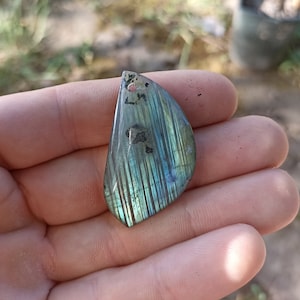 May include: A teardrop-shaped pendant made of iridescent labradorite with a green and blue color scheme. The stone has a striated pattern and a hole drilled at the top for hanging.