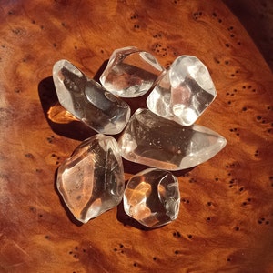 May include: A collection of six clear quartz crystals of various shapes and sizes, displayed on a brown wooden surface.
