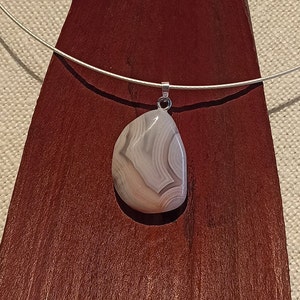May include: A pendant necklace featuring a teardrop-shaped stone with swirling patterns of gray and beige. The stone is suspended from a thin, silver-colored wire. The necklace is displayed on a dark red, wooden surface.