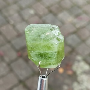 Peut inclure: Un cristal vert avec une surface brute et facetée. Le cristal est maintenu en place par un outil en métal.