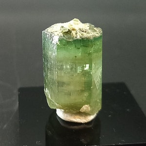 May include: A green and clear crystal with a hexagonal shape. The crystal is on a white base.
