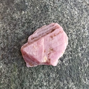 May include: A pink rough gemstone with brown inclusions. The gemstone has a rough, irregular shape and is a natural, unpolished crystal.