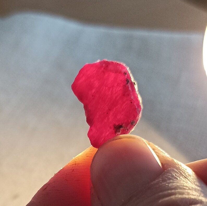 May include: A vivid, raw ruby gemstone, exhibiting a rich, deep red colour. The rough-cut stone has a textured surface with small dark inclusions. The ruby is held between fingers, showcasing its natural form and colour.